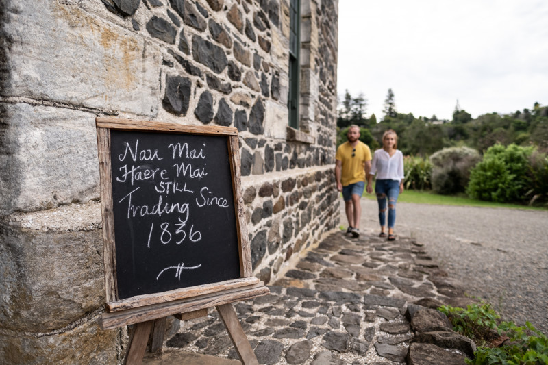 Couple walking along the path towards the Stone Store with an entry sign 'Nau Mai Haere Mai, Trading since 1836'