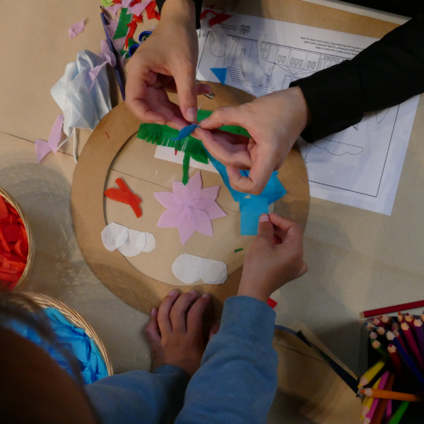 Four hands sticking coloured pieces of paper into a circular frame.
