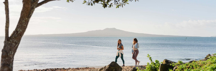 Women in athletic wear walking along Takapuna Beach early in the morning with Rangitoto in the background.
