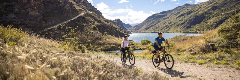 Couple cycling along rail trail with Lake Dunstan in the background